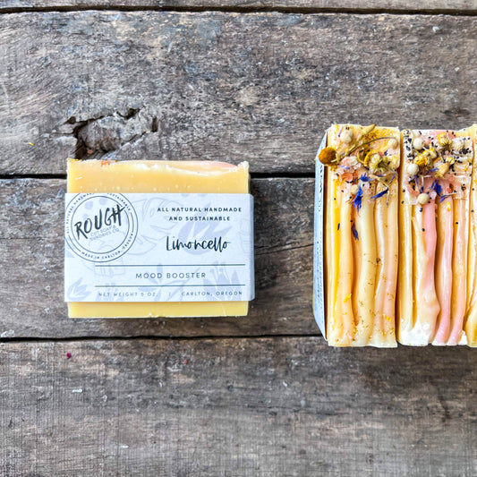 Two soap bars on a wooden surface, one with a label and the other with botanical toppings
