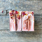 Three pink soap bars with rose petals on a wooden surface